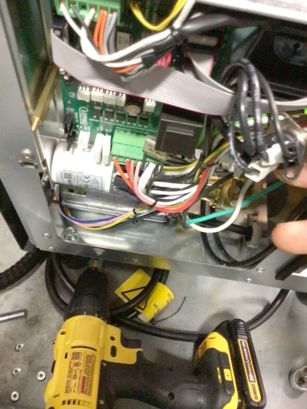 Close-up of an open electronic device showing various colored wires, circuit boards, and connectors. A person’s hand holds cables, and a yellow cordless drill is visible on the floor below the device. © 2025 1st-line.com