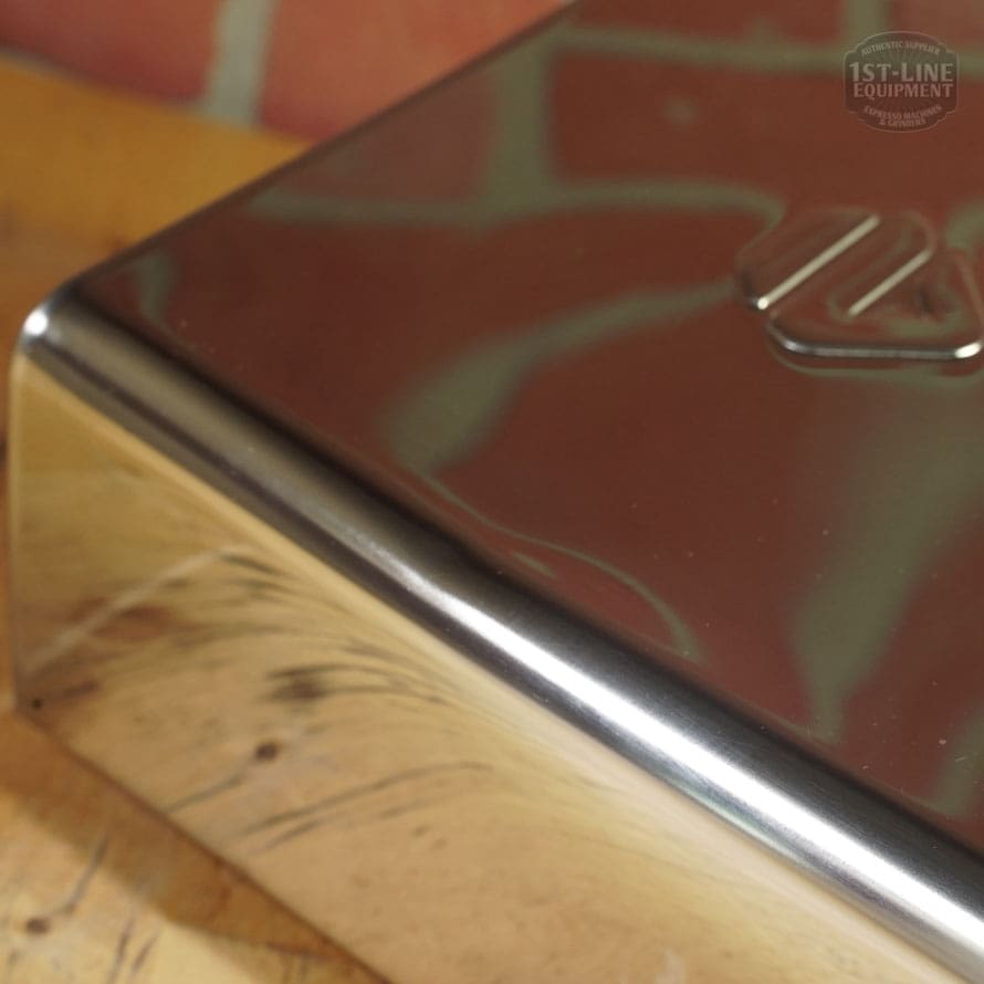 Close-up of the Lelit 1000156 External Water Tank Assembly - Small Dent & Scratch (E112) shows its shiny, reflective stainless steel corner with embossed logo, minor dents and scratches, on a wooden surface and brick wall background. © 2025 1st-line.com