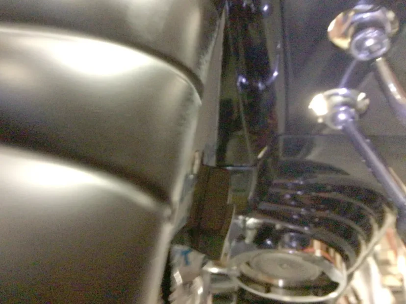 Close-up view of part of a motorcycle, showing the edge of a black leather seat, shiny metal bolts, and part of the engine with some components visible in the background. © 2025 1st-line.com