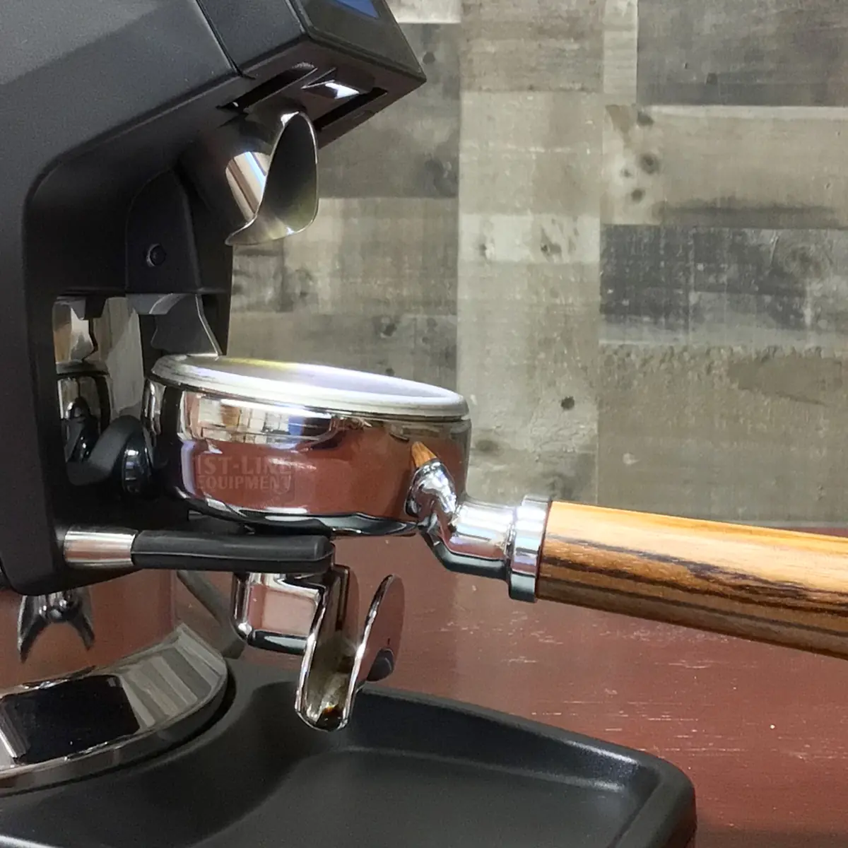 A close-up of a coffee machine with a portafilter attached, featuring a wooden handle and a shiny metal basket, ready for brewing espresso. The background shows a rustic wooden wall. © 2025 1st-line.com