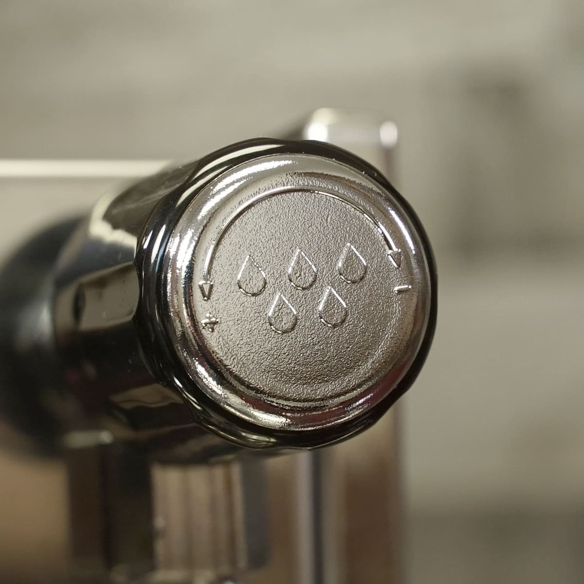 A close-up of a metallic espresso machine control knob featuring five droplet symbols and small directional arrows. The background is blurred. © 2025 1st-line.com