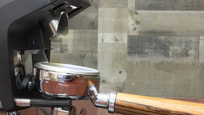 A close-up of a portafilter with a wooden handle attached to an espresso machine, set against a background of gray wooden panels. © 2025 1st-line.com