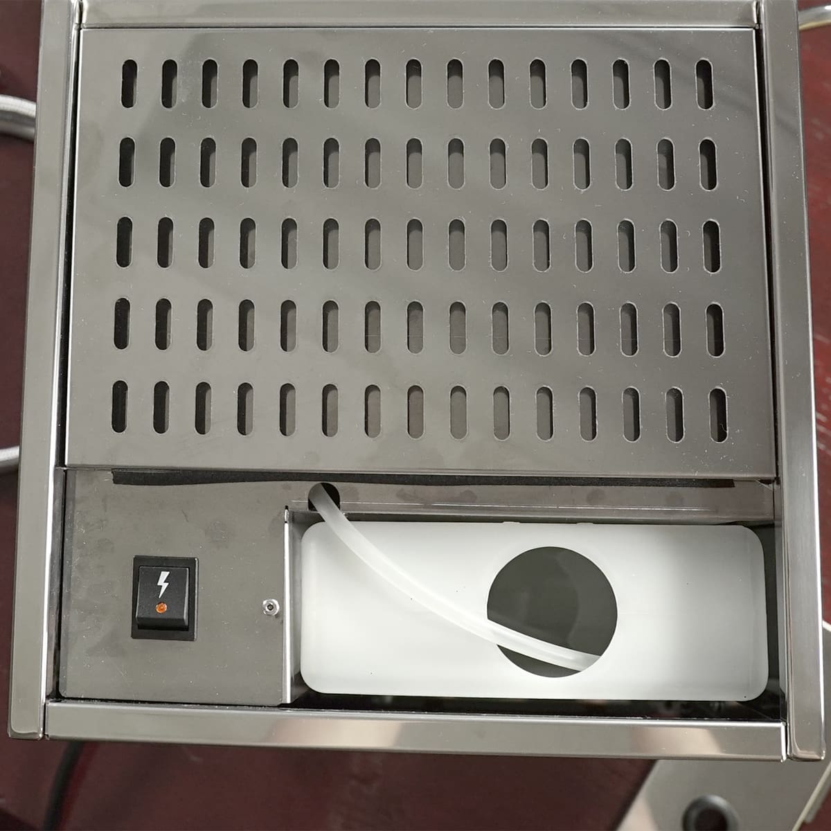 A close-up of a stainless steel appliance top with ventilation slots, a black power switch featuring a lightning bolt symbol, and a white plastic compartment with a tube and circular opening. © 2025 1st-line.com