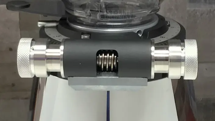 A close-up view of a coffee grinders adjustment mechanism, featuring metal knobs and a visible spring in the center, against a blurred background. © 2025 1st-line.com