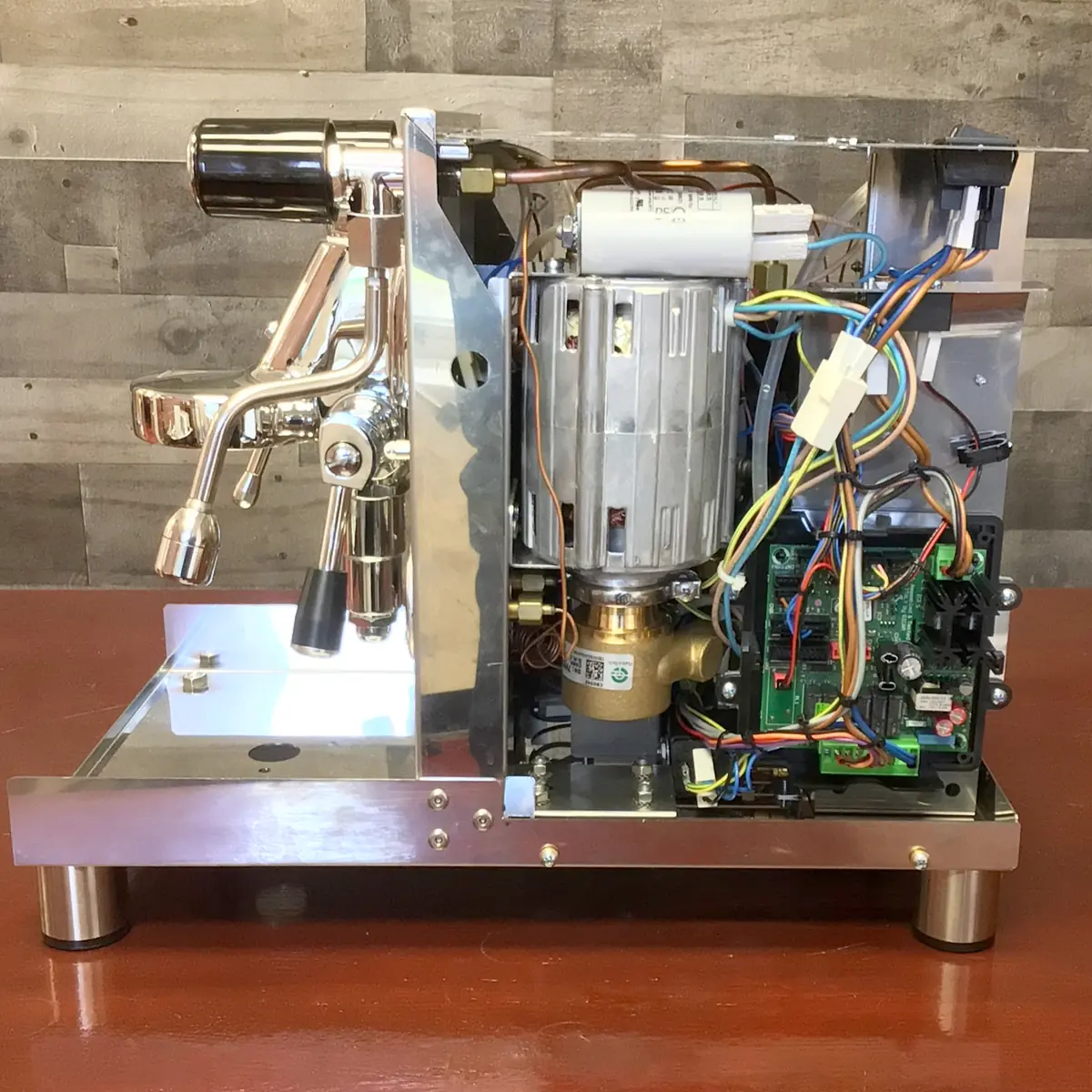 A partially open espresso machine showing internal components like wires, a motor, circuit boards, and pipes, on a wooden table with a wood-paneled wall background. © 2025 1st-line.com