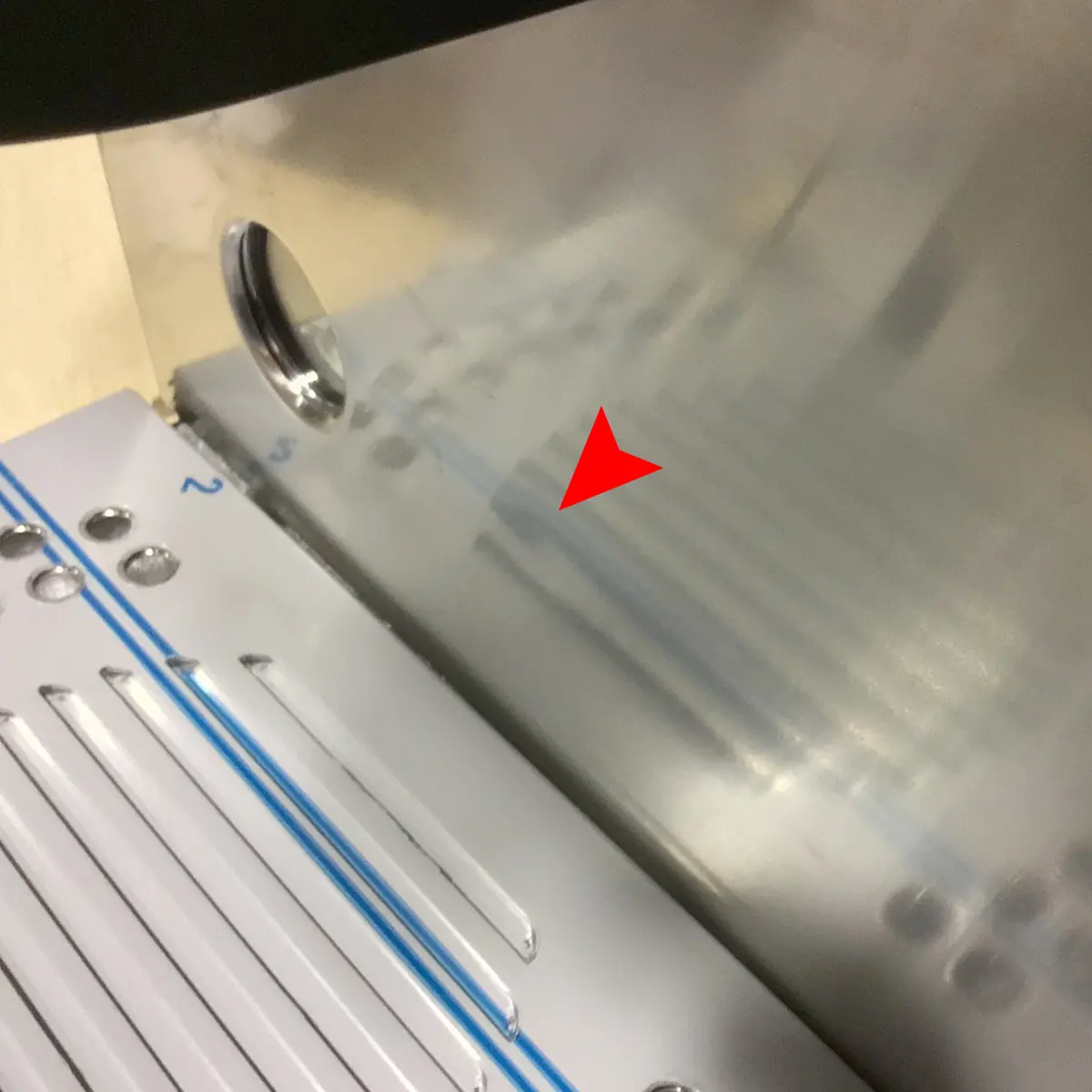 A red arrow points to faint blue marks or streaks on a shiny metallic surface next to a white panel with blue lines and metal screws. © 2025 1st-line.com