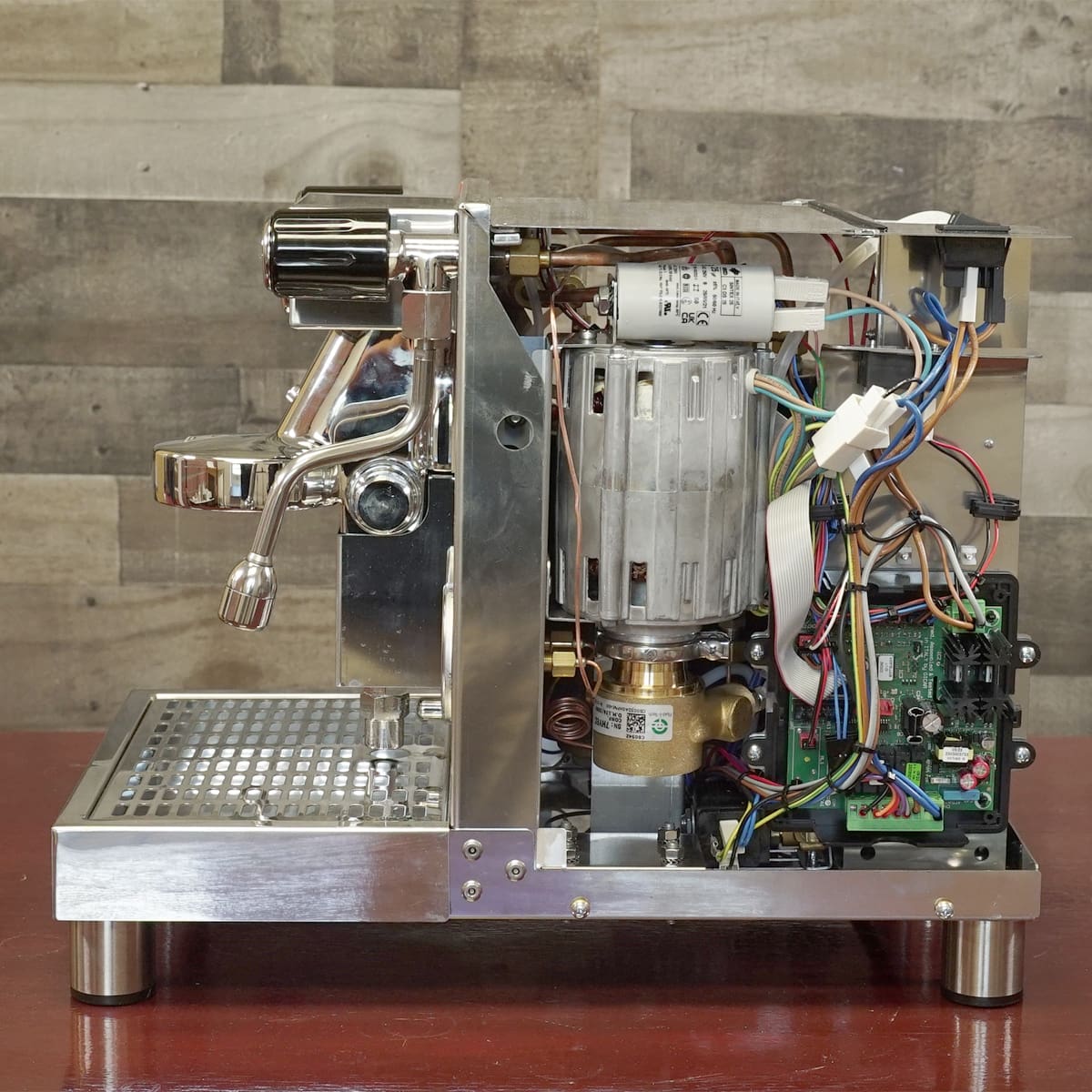 A semi-automatic espresso machine with its side panel removed, exposing internal components like wiring, the pump, circuit board, and metal framework, sitting on a brown surface against a wood-patterned wall. © 2025 1st-line.com