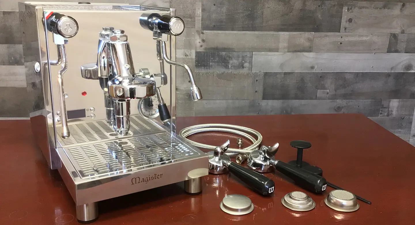 A shiny stainless steel espresso machine sits on a brown surface next to accessories including portafilters, a tamper, metal filter baskets, and hoses. The machine has dials, a steam wand, and is labeled Magister. © 2025 1st-line.com