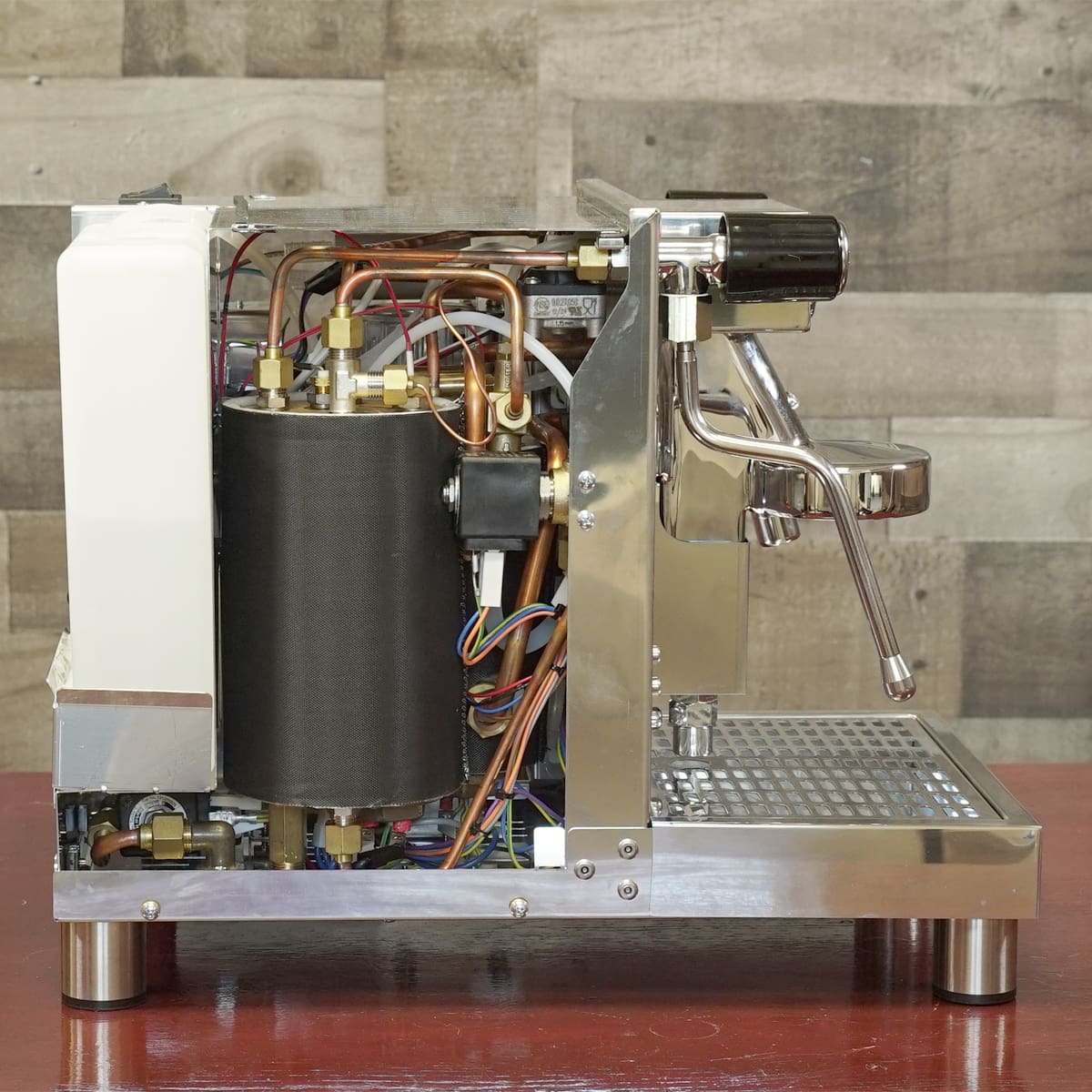 A side view of an espresso machine with its cover removed, revealing internal components like wires, pipes, a boiler, and metal framework, displayed on a red surface against a wooden panel background. © 2025 1st-line.com