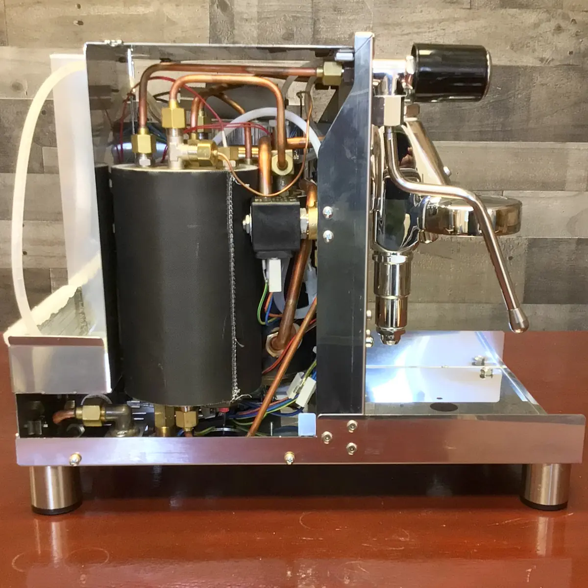 A side view of an espresso machine with its outer casing removed, revealing internal components such as pipes, wires, and a boiler against a wooden background. © 2025 1st-line.com