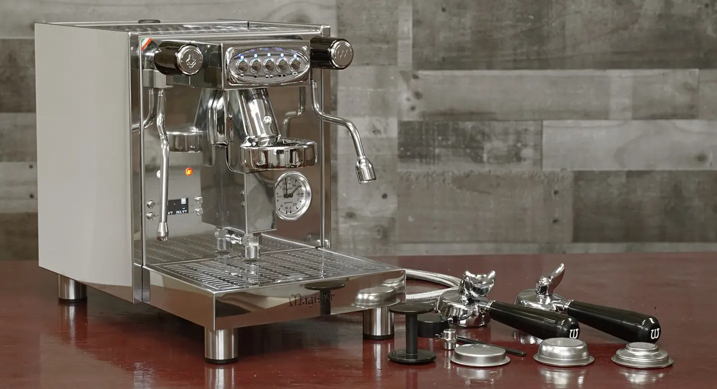 A stainless steel espresso machine sits on a brown table with various accessories, including portafilters, a tamper, and baskets, arranged beside it. A textured gray wall is in the background. © 2025 1st-line.com