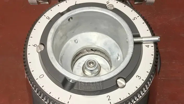 Close-up of a coffee grinder burr assembly with numbered calibration dial, showing the central burr, adjustment mechanism, and metal parts on a brown surface. © 2025 1st-line.com