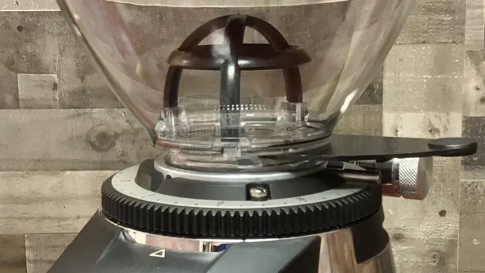Close-up of a coffee grinder hopper and adjustment dial with a textured, wooden background. The clear plastic hopper sits atop the metal grinder, showing a black internal component and part of the grind size adjustment ring. © 2025 1st-line.com