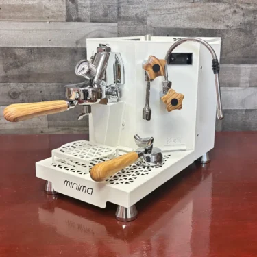 A white espresso machine with the Minima Walnut Wood Kit—wood handles and knobs—sits on a reddish-brown countertop, featuring a steam wand, pressure gauge, and distinct minima branding on the front. © 2025 1st-line.com