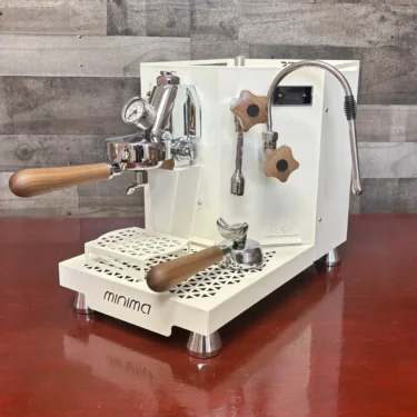A Minima Walnut Wood Kit espresso machine with walnut handles, metal steam wand, pressure gauge, and portafilter rests on a reddish-brown surface before a textured wood panel backdrop. © 2025 1st-line.com