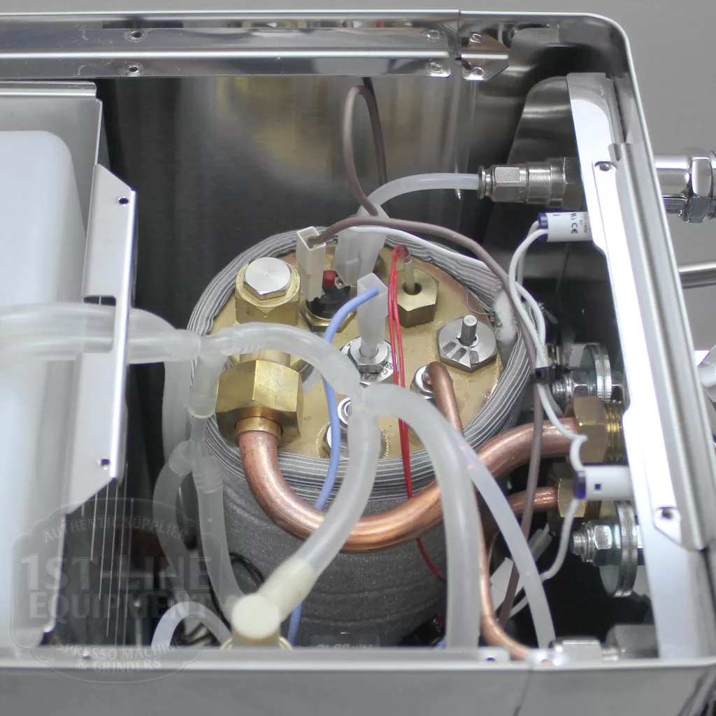 Close-up view of the internal components of an espresso machine, showing a metal boiler with copper pipes, wires, and connectors inside a stainless steel casing. © 2025 1st-line.com