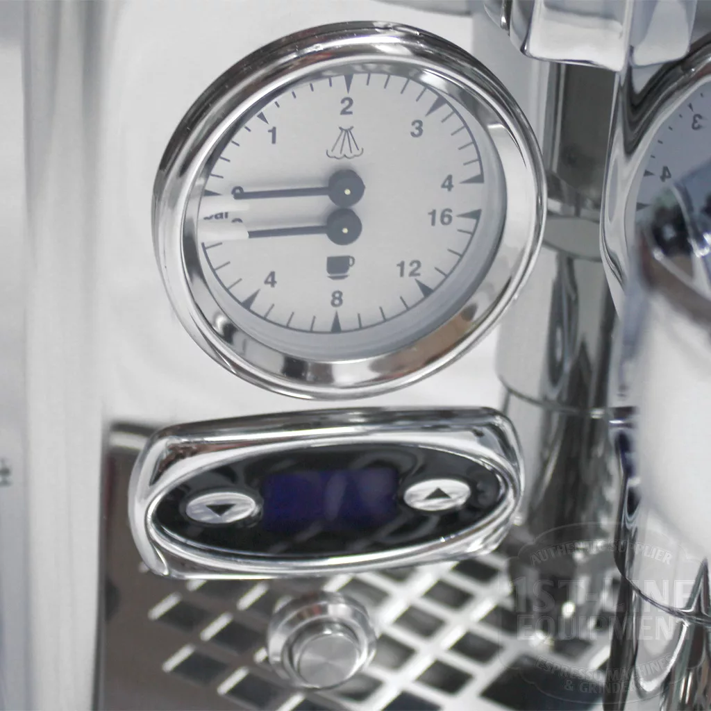 Close-up of a shiny metal gauge with black markings and a needle, mounted on a stainless steel espresso machine, above a drip tray with a button and digital display below. © 2025 1st-line.com