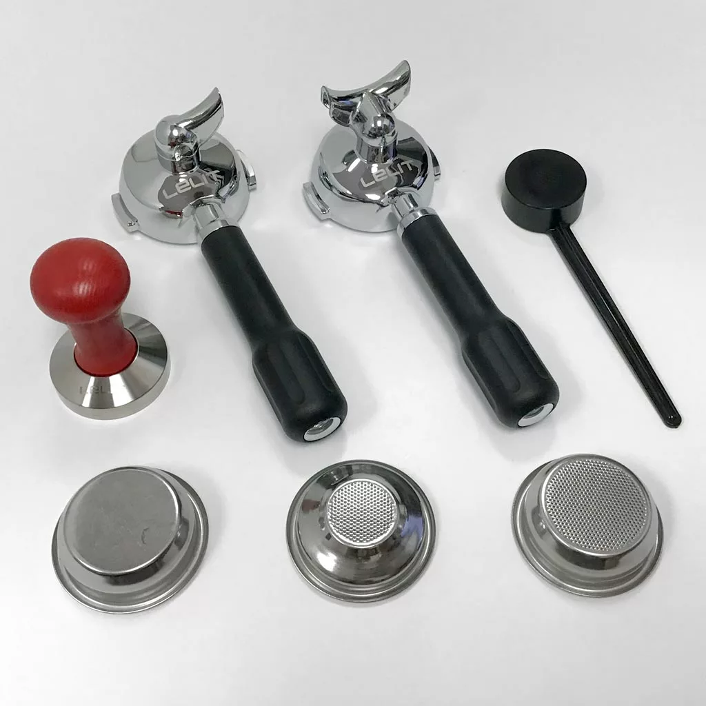 Assorted espresso machine accessories on a white surface, including two portafilters with black handles, a red-handled tamper, a black dosing tool, and various metal filter baskets and inserts. © 2025 1st-line.com