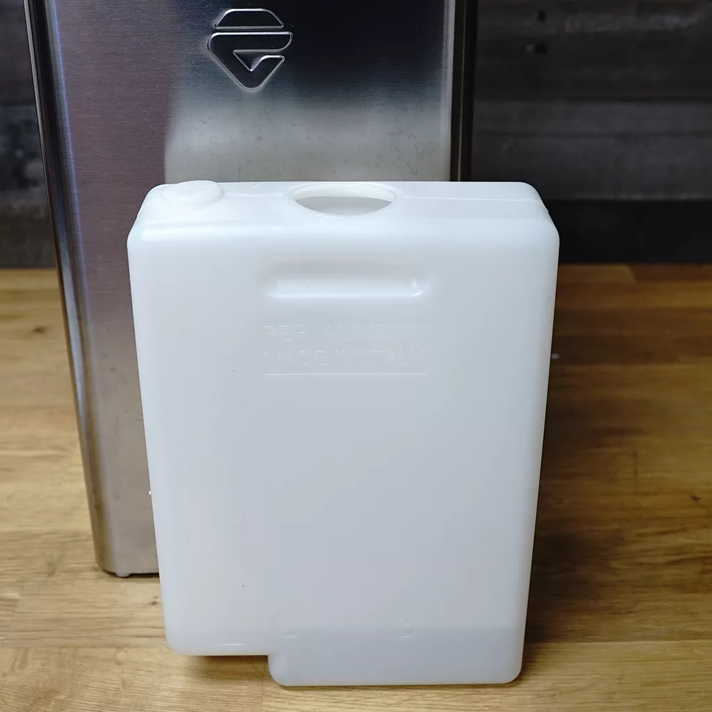 A white rectangular plastic water reservoir sits on a wooden surface in front of a stainless steel appliance, with a circular opening visible on top. &copy; 2025 1st-line.com