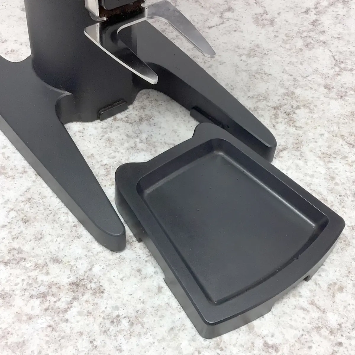 A close-up of a black plastic base with a detachable drip tray on a speckled countertop, part of the SIDEWALK SALE Pinecone Pinion All-Purpose Espresso Coffee Grinder (showroom demo). © 2025 1st-line.com