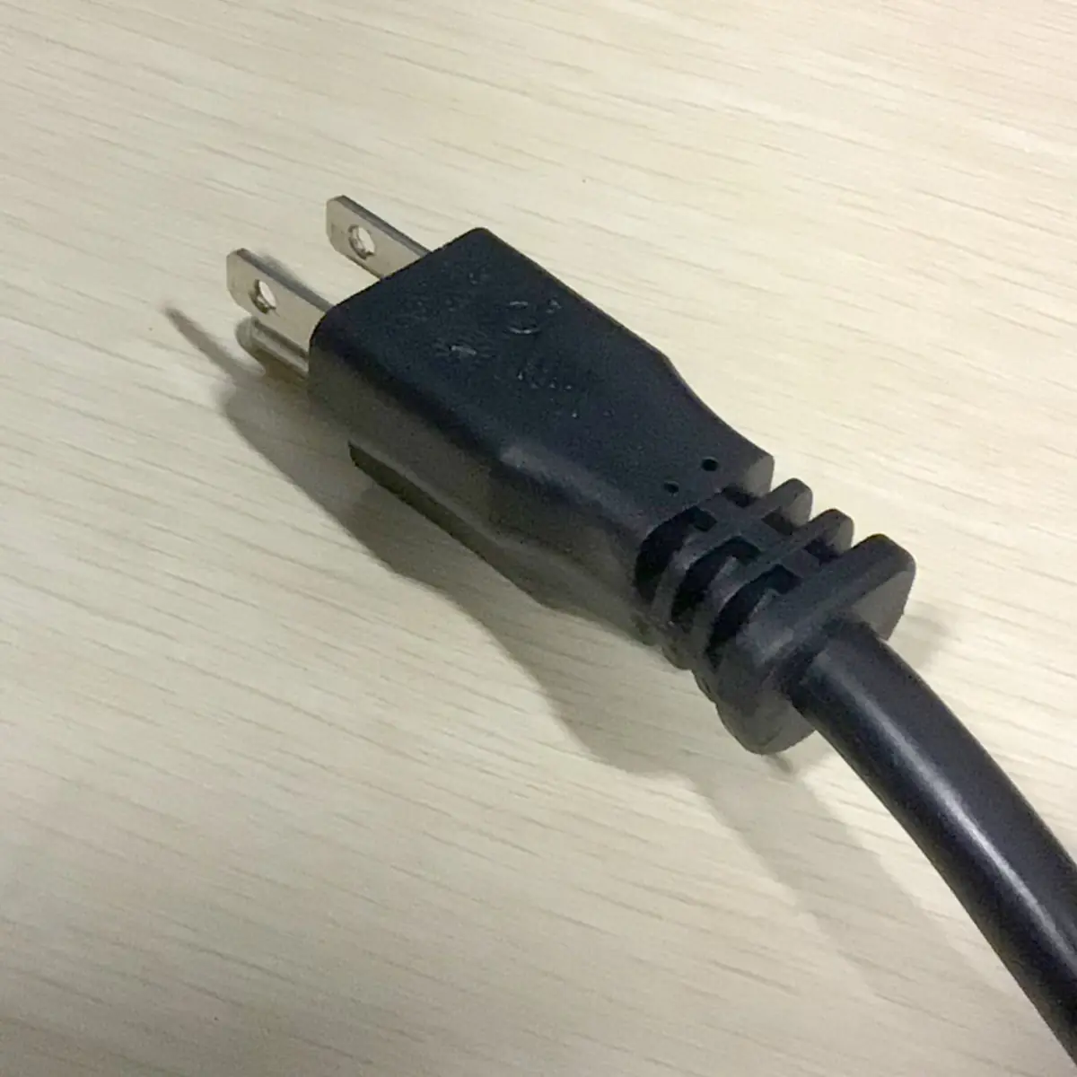 A close-up of a black, two-prong electrical power plug resting on a light-colored wooden surface. © 2025 1st-line.com