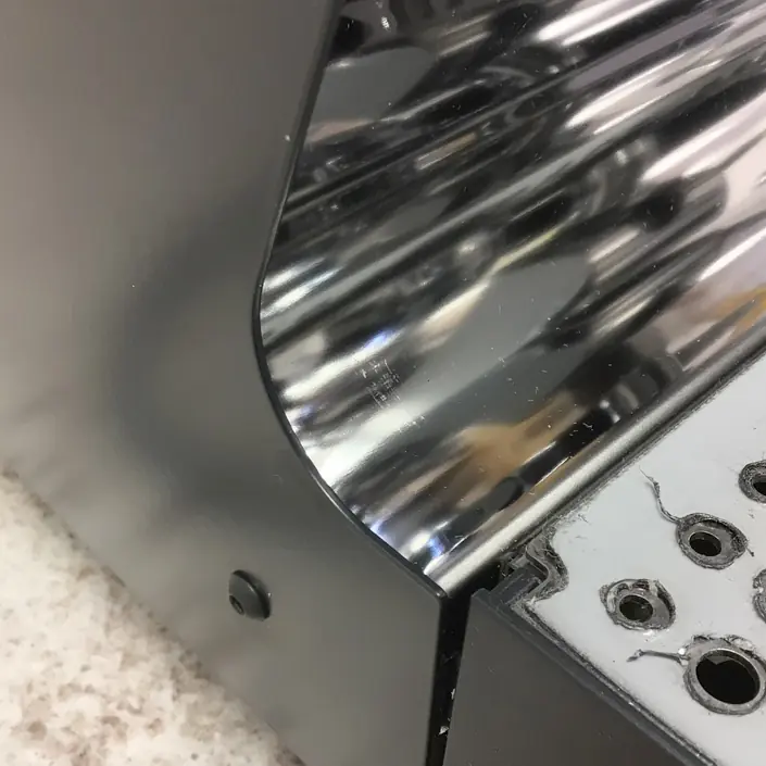 A close-up of a metallic, reflective surface with curved ridges next to a section with circular holes, attached to a textured, light-colored countertop. © 2025 1st-line.com