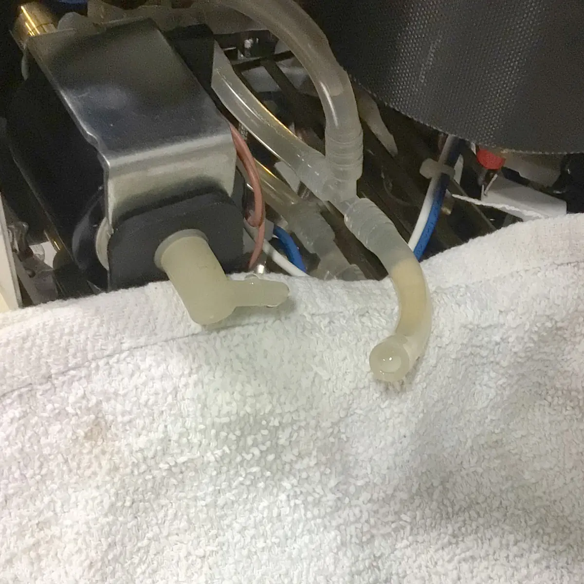 A close-up of disconnected transparent tubing and a plastic nozzle on a machine, partially covered by a white towel. Various wires and components are visible in the background. © 2025 1st-line.com