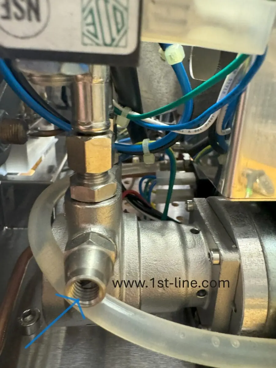 A close-up of metallic plumbing parts with wires and a clear tube attached, highlighting a threaded port with a blue arrow, and the website www.1st-line.com visible on the metal surface. © 2025 1st-line.com