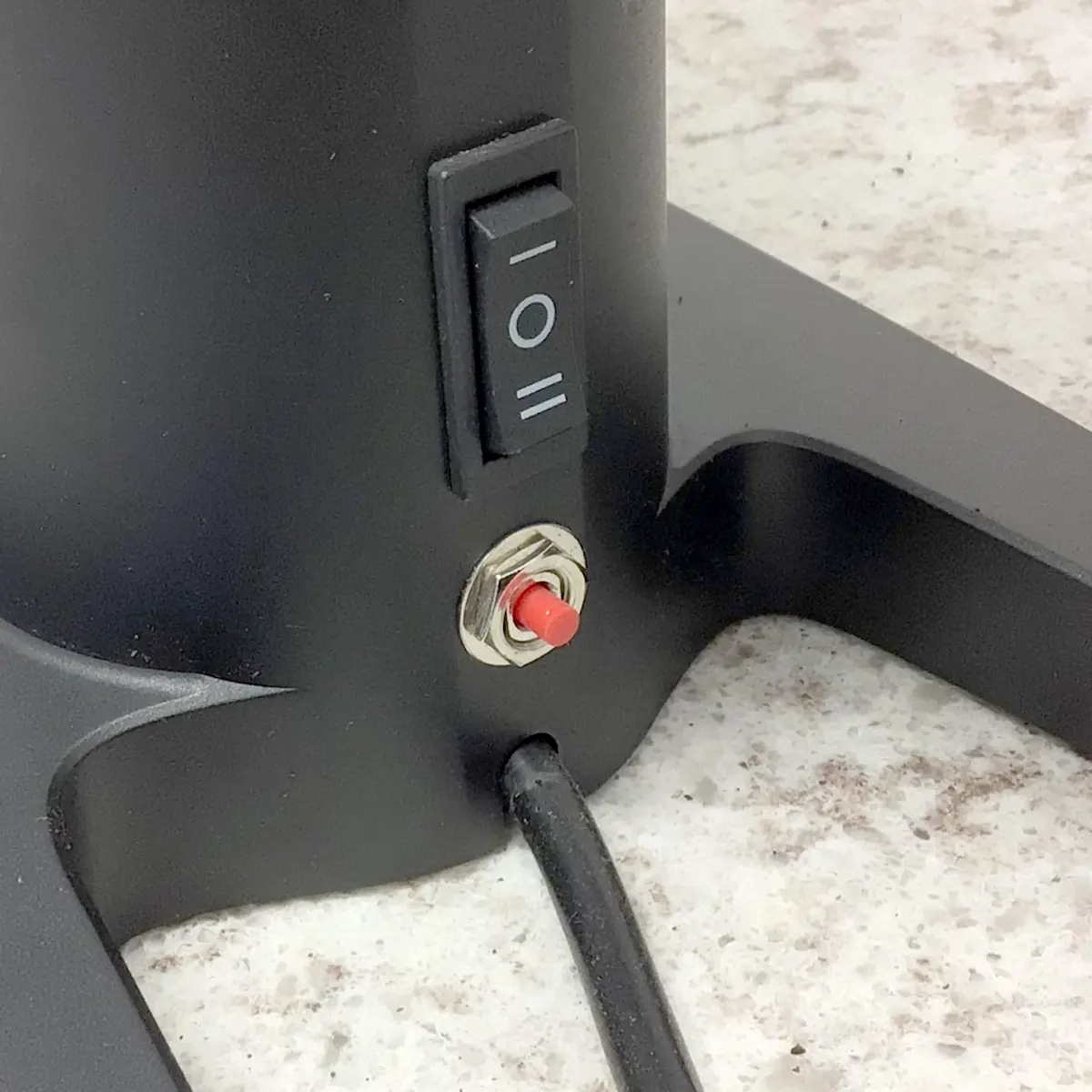 A close-up of the SIDEWALK SALE Pinecone Pinion All-Purpose Espresso Coffee Grinder (showroom demo) base shows an on/off rocker switch with I/O symbols, a red push button, and a black power cord on a speckled surface. © 2025 1st-line.com