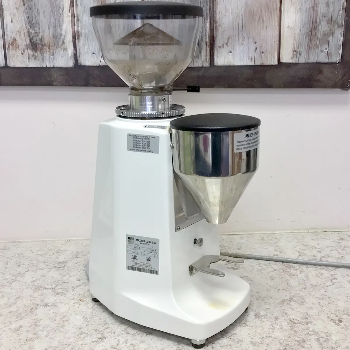 A commercial coffee grinder with a white base and two transparent hoppers sits on a speckled countertop against a rustic wooden wall. The machine has labels and a power cord visible on the side. © 2025 1st-line.com