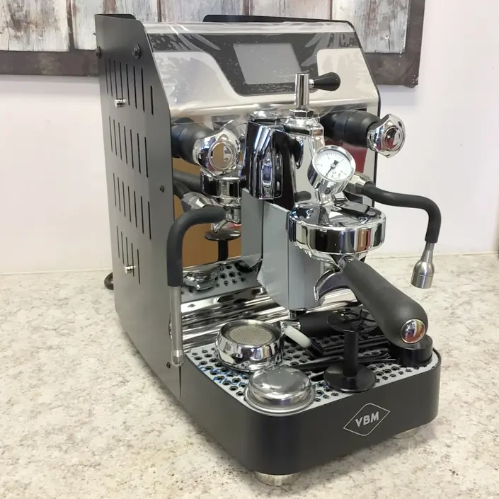 A shiny, stainless steel espresso machine with black accents sits on a light-colored countertop. The machine features a pressure gauge, steam wand, portafilter, and the VBM logo on the front. © 2025 1st-line.com