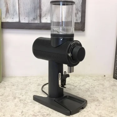 A SIDEWALK SALE Pinecone Pinion All-Purpose Espresso Coffee Grinder (showroom demo) with a transparent bean hopper is on a light countertop, set against a white wall and wooden panel. © 2025 1st-line.com