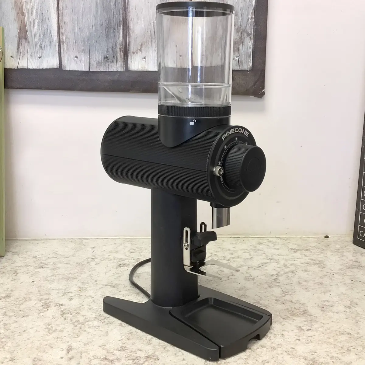 A SIDEWALK SALE Pinecone Pinion All-Purpose Espresso Coffee Grinder (showroom demo) with a transparent bean hopper is on a light countertop, set against a white wall and wooden panel. © 2025 1st-line.com