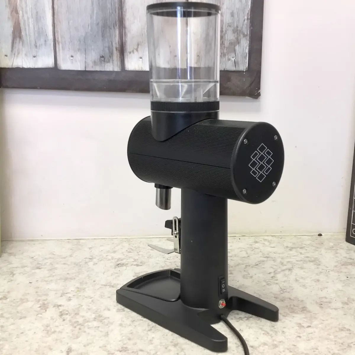 A SIDEWALK SALE Pinecone Pinion All-Purpose Espresso Coffee Grinder (showroom demo) with a transparent hopper is displayed on a speckled countertop in front of a rustic wooden wall panel. © 2025 1st-line.com