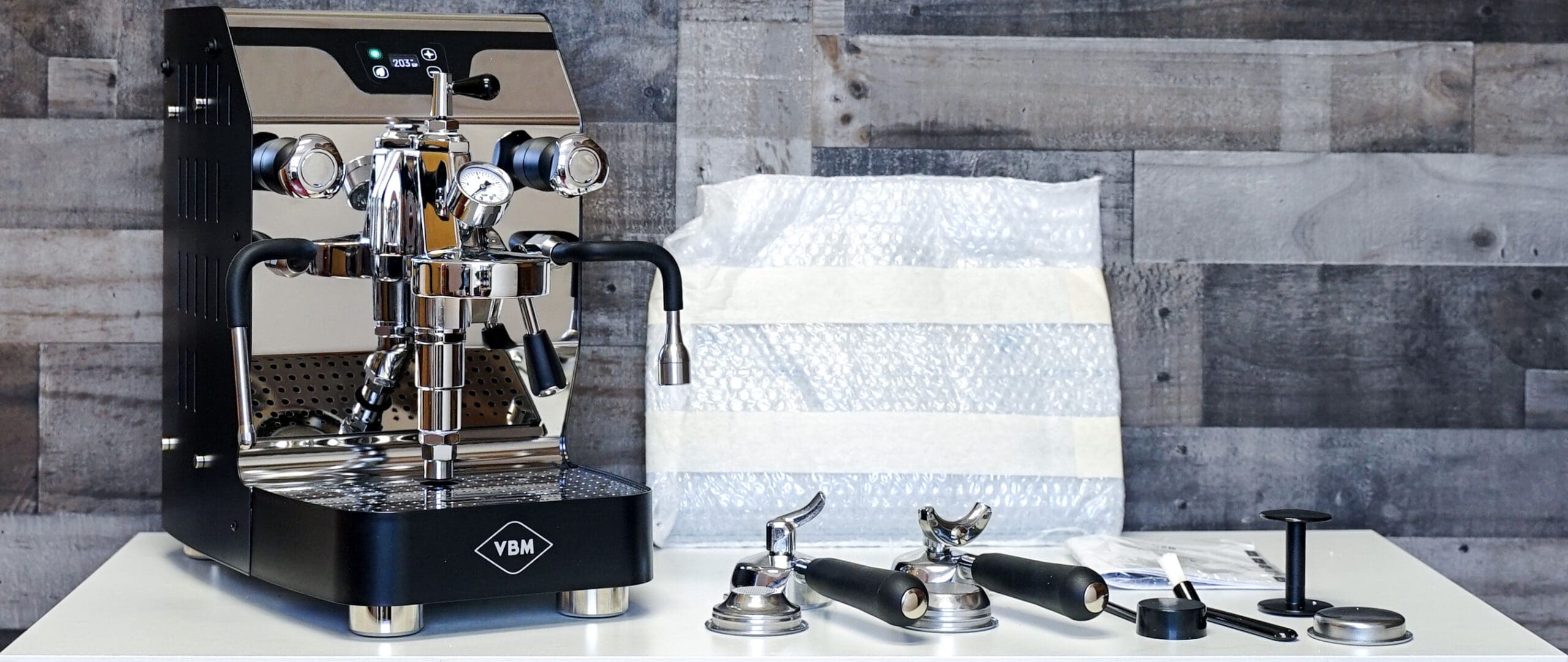 A stainless steel espresso machine with gauges and two levers sits on a white counter, surrounded by portafilters, a tamper, and a packing material background. © 2025 1st-line.com