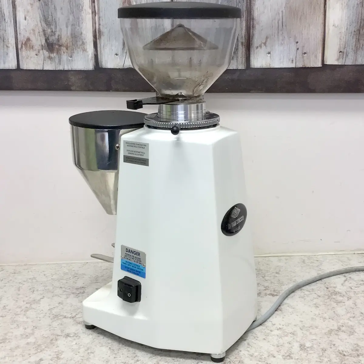 A white electric coffee grinder with a transparent hopper sits on a light-colored countertop against a wooden wall background. The grinder has a black power switch and a visible power cord. © 2025 1st-line.com