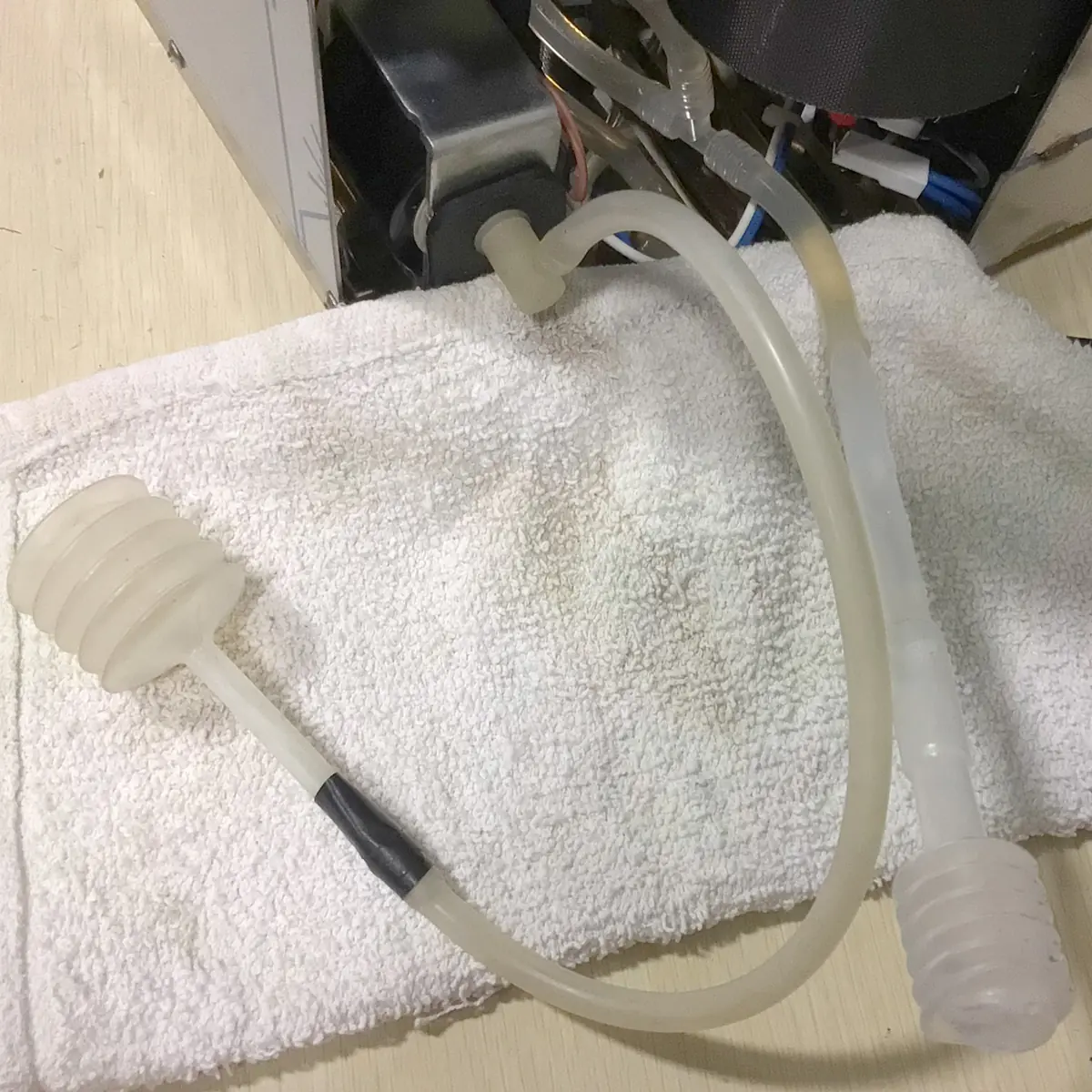 A white towel with a clear flexible tube on top, featuring two ribbed, cylindrical silicone ends. The tube is partially connected to an open machine with visible wires and internal components. © 2025 1st-line.com