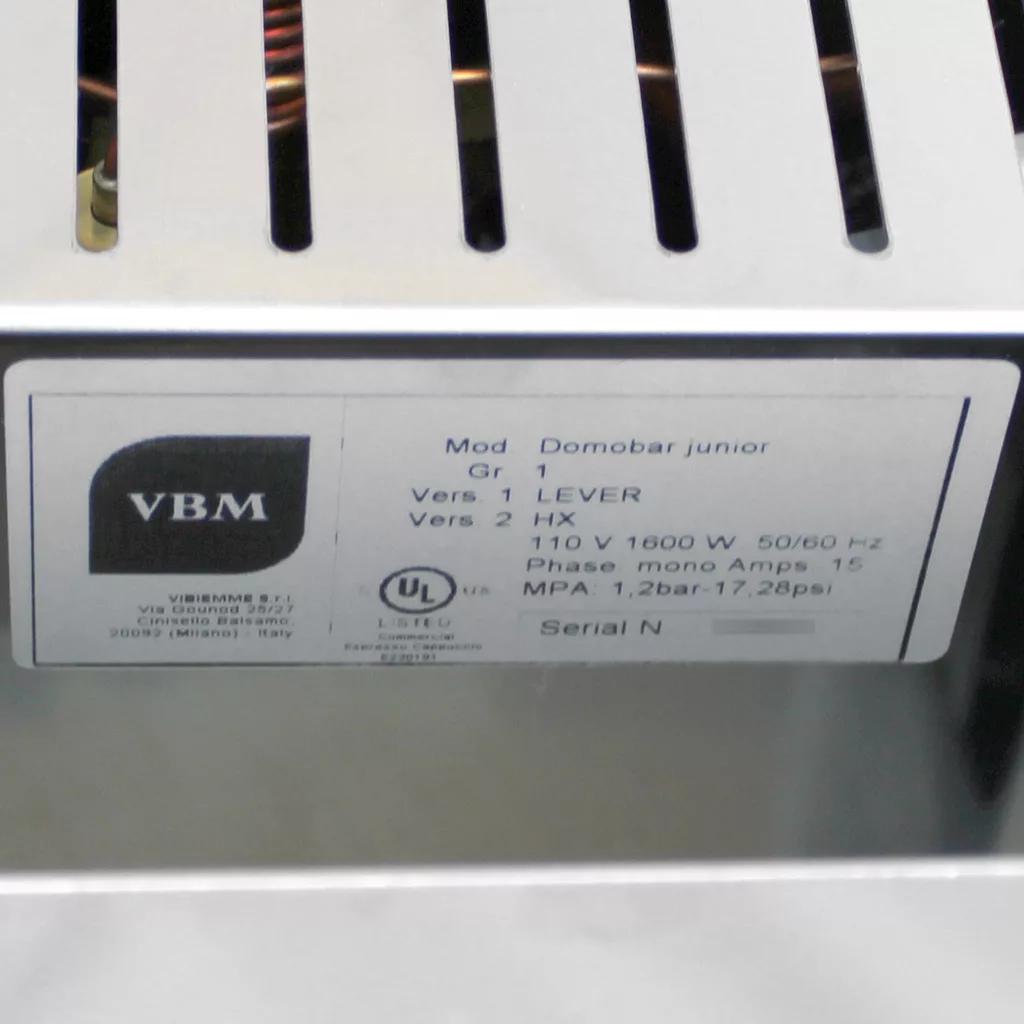 Close-up of a metal label on a VBM Domobar Junior espresso machine showing technical specifications including voltage, wattage, frequency, phase, and pressure details. The label also includes certification logos. © 2025 1st-line.com