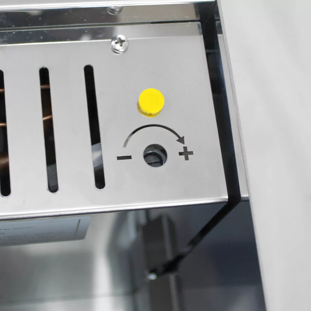 A close-up of a stainless steel control panel shows a yellow button above a circular dial slot with plus and minus symbols and a curved arrow indicating adjustable settings. © 2025 1st-line.com