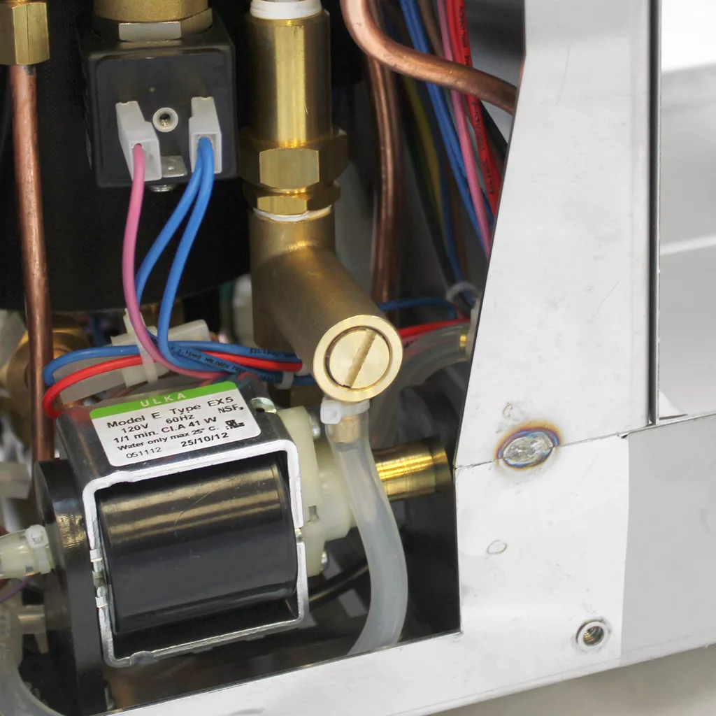 Close-up view of internal components of a machine, including wires, copper pipes, a brass fitting with a slot for a screwdriver, and a small electronic part labeled “Model Type EX5.”. © 2025 1st-line.com