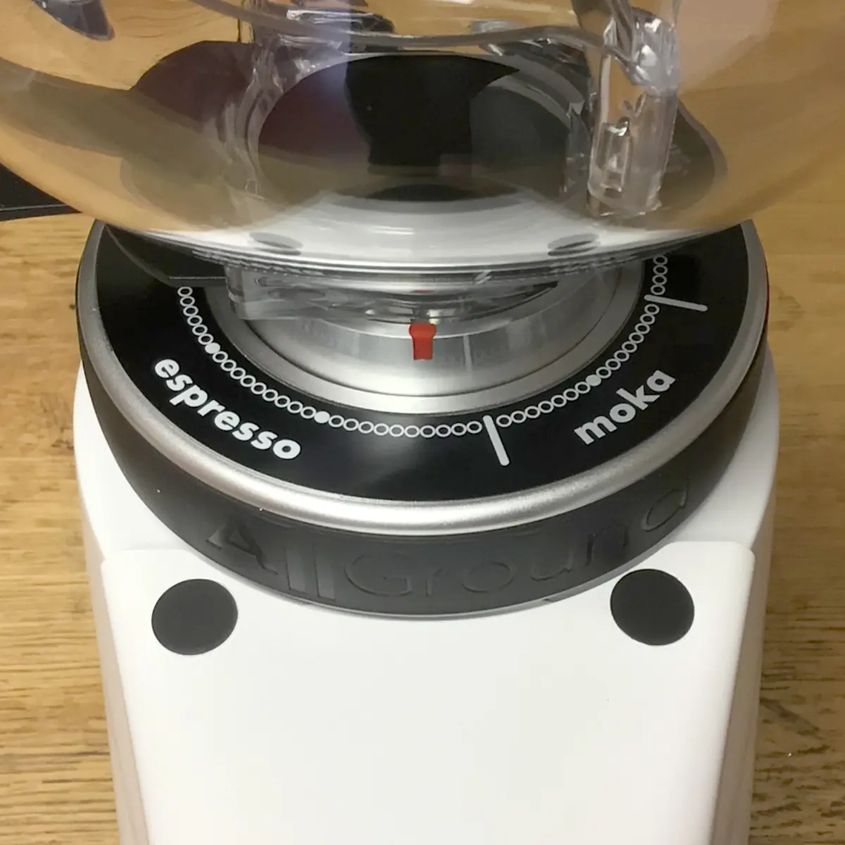 Close-up of a coffee grinder dial with espresso and moka grind settings, a red indicator, and the word AllGround embossed on the white grinder base. The surface underneath is light wood. © 2025 1st-line.com
