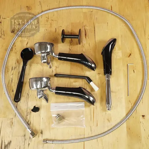 Espresso machine accessories on a wooden surface, including a portafilter, steam wand, group head brush, tamper, spoon, hose, cleaning tool, and small parts in a plastic bag. © 2025 1st-line.com