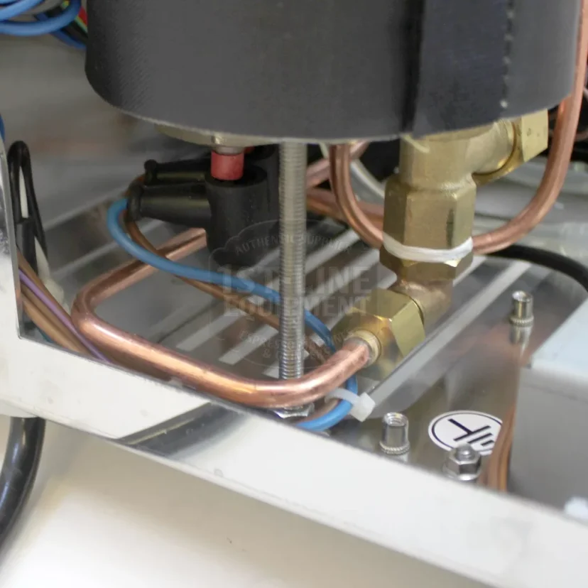 Close-up of copper pipes, brass fittings, blue wires, and a metal rod inside a metal machinery casing, likely part of industrial or coffee machine equipment. © 2025 1st-line.com