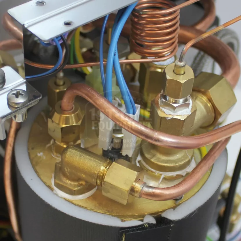 Close-up of a metal component featuring copper pipes, brass fittings, and coiled copper tubing connected to a cylindrical base with wires and bolts, part of a mechanical or electrical machine. © 2025 1st-line.com