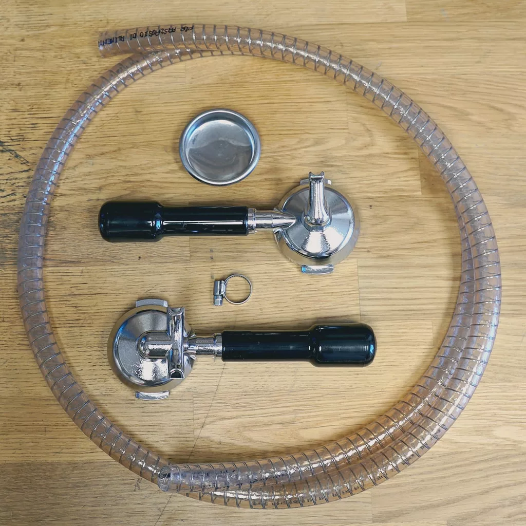 Two portafilters with black handles, a tamper, a metal hose clamp, and a coiled clear plastic hose are arranged on a light wooden surface. © 2025 1st-line.com