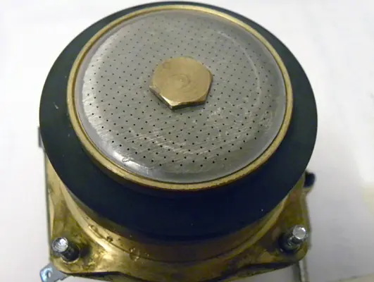 Close-up of a metal surface with small holes, a central hexagonal bolt, and a round black rim, likely part of a Lelit PL41 espresso machine's Dispersion Screen Upgrade or similar mechanical device. 2025 1st-line.com