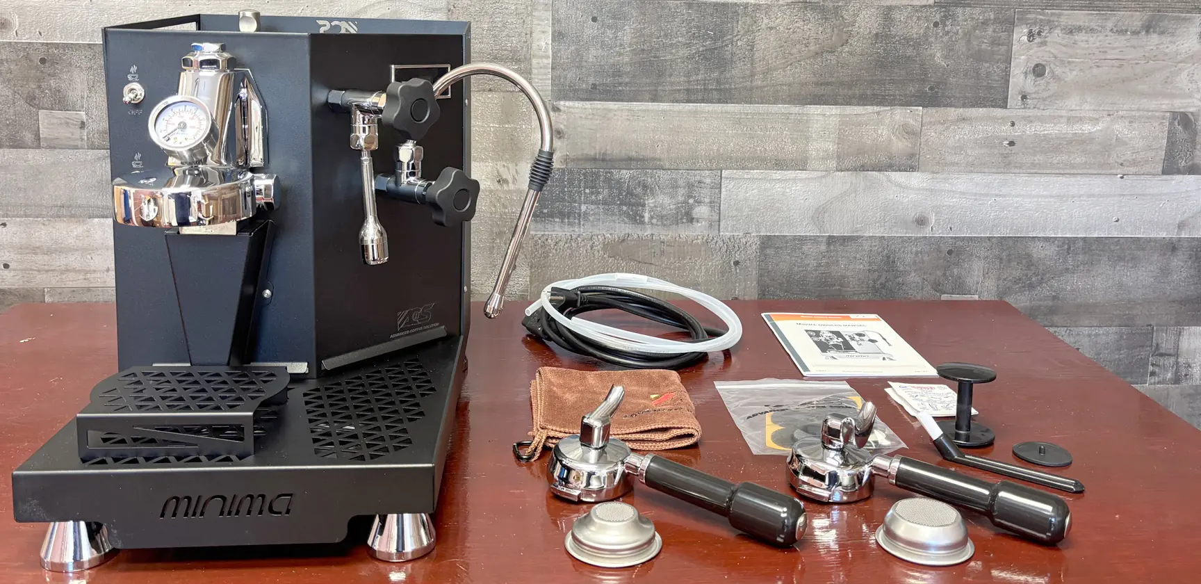 A black espresso machine sits on a wooden table next to accessories, including portafilters, baskets, a tamper, a cloth, cables, an instruction manual, and other small parts. The background is a wood-paneled wall. © 2025 1st-line.com