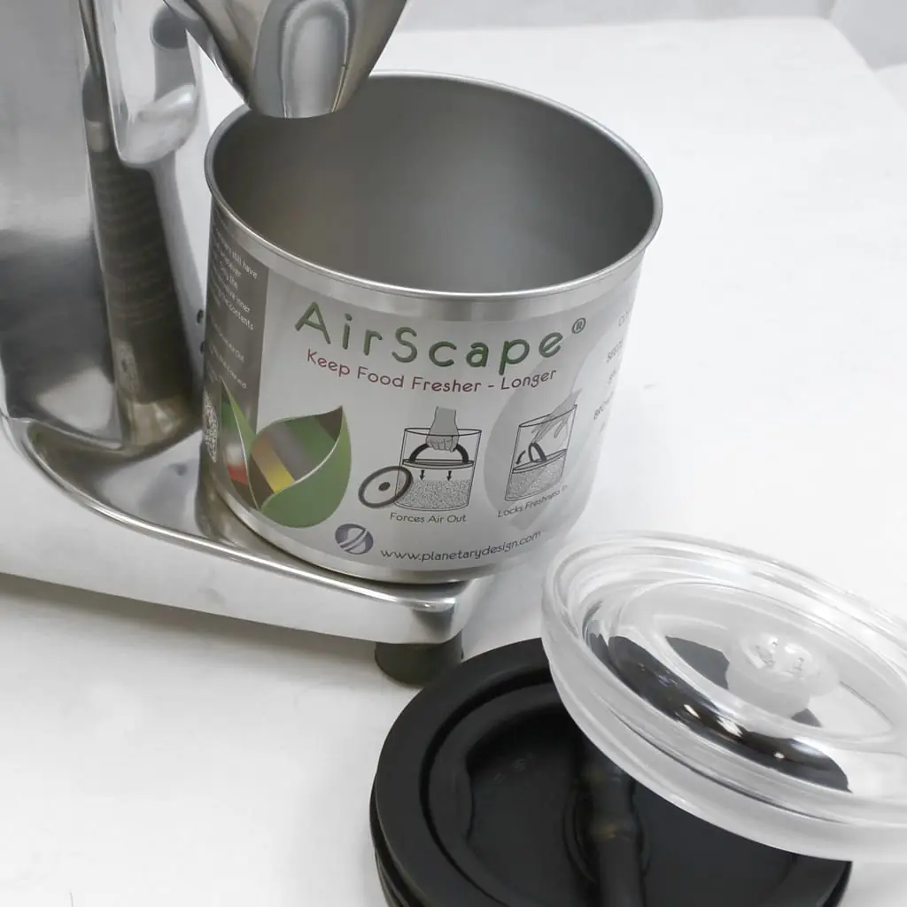 A stainless steel AirScape food storage canister sits open on a counter next to its clear plastic and black lids, with part of a Q50 metal appliance visible beside it. 2025 1st-line.com