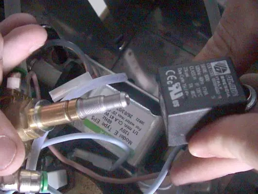 Close-up of hands holding an electrical solenoid valve component, ideal for Solenoid Valve Repair on machines like the Lelit PL41 Anna, with visible wires and hardware in the background. The solenoid shows its specifications and certification markings. 2025 1st-line.com