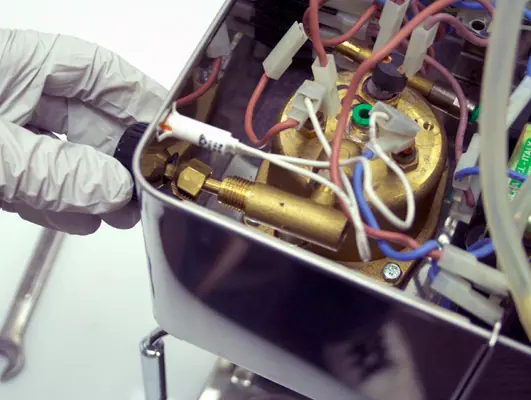 A gloved hand adjusts a component inside an open Lelit PL41 Anna machine filled with wires and brass parts, revealing its internal mechanisms during a steam valve repair. 2025 1st-line.com